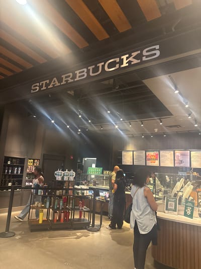 Here is a detailed description of the image:
**Scene:** Interior of a Starbucks coffee shop. There are customers waiting in line to order and purchase items.
**Environment:** The Starbucks is well-lit with track lighting. There's a menu board above the counter. The floor appears to be concrete, and there's a display of merchandise including cups and packaged snacks.
**People:** There are several people, customers, lining up and potentially placing orders at the counter. One employee is visible behind the counter.
**Objects:**
* Starbucks sign
* Menu board
* Display of cups
* Order counter
* A sign with a QR code
* Cups and supplies behind the counter.
**Actions:** People are waiting in line, examining the merchandise, and interacting with the barista.
**Interactions:** Customers are waiting and potentially ordering.
**Location**
The image shows the interior of a Starbucks coffee shop. Without street signs or other details, it is difficult to determine the exact location within the city of Chicago, United States.