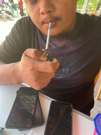 A male individual is observed in Sagoe, Indonesia, within an outdoor or semi-outdoor setting during daylight hours. The subject, visible from the chest upwards, has dark hair, stubble, and a mustache, and is wearing a black t-shirt with a dark logo. He is actively lighting a cigarette held between his lips using a metallic lighter with a green detail in his right hand; a flame is visible at the cigarette tip. The cigarette is white with a light blue and yellow band near the filter. On a white table in the foreground, two black smartphones are present. The left phone displays a Wi-Fi settings screen showing "OPPO A1s nbga" and the time "15:35." The right phone has a dark screen with minor visible damage to its upper right corner. Partially visible items on the table include a small white packet and a blue and white container. The blurred background shows motorcycles, other individuals, green foliage, and light-colored structures, with a yellow chair leg visible on the far right.