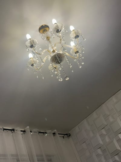 This image captures a static interior scene in Marcoussis, France. The primary subject is an ornate crystal chandelier, centrally positioned and suspended from a flat, white ceiling. The chandelier features a gold-toned metallic frame with multiple arms extending outward, supporting at least eight illuminated, candle-style light bulbs. Each bulb is encased in a clear glass or crystal cup, and numerous transparent crystal pendants, varying in shape from teardrops to small spheres, dangle from the chandelier's structure. Below, in the lower left, a sheer white curtain is visible, hung from a black metal curtain rod with a decorative finial on the right. To the lower right, a portion of a white wall is visible, textured with a repeating three-dimensional pattern of raised geometric shapes, likely squares or rectangles. Faint light reflections are discernible on the surrounding white surfaces, emanating from the chandelier's illuminated crystals. No individuals or active subjects are present.