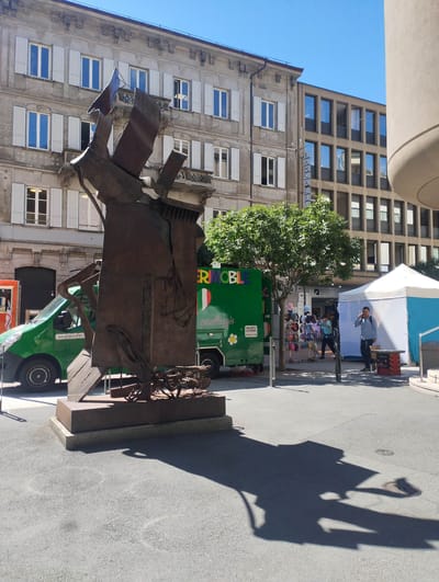 This image captures a vibrant urban scene in Bellinzona, Switzerland, under clear, bright daylight. The central subject is a large, dark, abstract metal sculpture featuring angular and fragmented forms, casting long shadows on the asphalt ground, suggesting midday or early afternoon.
In the background, two distinct architectural styles are visible: an older, light-colored multi-story building with numerous windows featuring white shutters, and a more modern, darker building with a grid-like facade of rectangular windows.
Several subjects populate the scene. To the left of the sculpture, a green commercial van with "www.pizzocherli.ch" visible on its side is parked. Behind the sculpture, a larger green commercial vehicle or food truck is present, displaying "ERIMOBILE," "gemellaggio," an Italian flag symbol, "SERVIZIO CATERING," and a phone number (079 387 67 26). A small tree stands between this vehicle and the tents.
On the right side of the frame, a white and blue temporary tent is set up. Near it, a man in a blue shirt is seen talking on a mobile phone, holding a small bag. Further back, another person is visible near a stand displaying merchandise, possibly hats or other accessories. The visible text on the modern building includes "BANCA DEL," likely part of a bank's signage.
The scene depicts an active outdoor environment, possibly a market or event space, with people moving and gathering amidst the architecture and commercial setups. The clear blue sky and strong shadows indicate sunny weather.