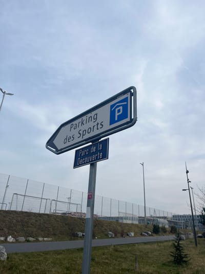 The image displays an outdoor scene, rotated 90 degrees clockwise, predominantly featuring a signpost with two signs under an overcast sky. In the foreground, a large white rectangular sign with a blue "P" parking symbol clearly indicates "Parking des Sports" in vertical black lettering, with an arrow pointing left. A smaller, darker blue rectangular sign below it reads "Parc de la Découverte" in white text. A small "CLEVER" label is visible on the upper left edge of the main sign's frame.

The signpost is metal and stands beside a grassy area with sparse rocks and a paved path running parallel to a low, transparent or mesh fence structure. Beyond the fence, there's more grass and slightly elevated ground. Several tall, slender utility poles or lampposts are visible in the middle ground and background, some with light fixtures. Distant buildings can be discerned on the horizon, suggesting an urban or suburban setting. The sky is uniformly gray and cloudy, indicating an overcast day. No people or specific activities are present, giving the scene a quiet and functional appearance. The French text on the signs confirms the context of Meyrin, Switzerland, provided.