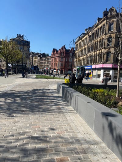 The image shows a cobbled street in Bradford, UK, located at 53.793° N, 1.753° W. The street is lined with buildings on both sides. There is a large, modern, gray stone planter on the right side of the image, separating a cobblestone sidewalk from a grassy area. Two men are sitting on the planter. The sidewalk is empty and there are several people walking in the street in the background. The day is sunny and clear, and the sky is blue. There are no cars present in the image. The buildings are primarily brick and stone, and have windows on all sides. The buildings on the left side of the image are relatively new and modern. The buildings on the right side of the image appear to be older. The pavement is made of gray cobblestones and the walls of the planter are made of smooth, gray stone. Trees are present in the image, but they are mostly bare-branched. The image is taken from a low angle.