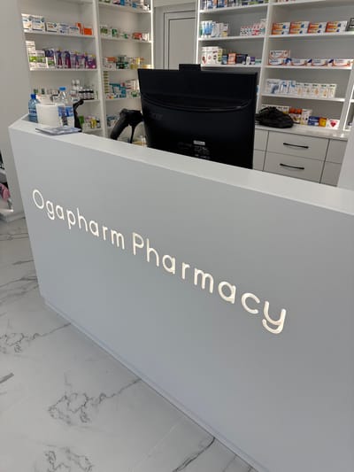 The image shows the interior of a pharmacy. The main subject is a white counter with the words "Ogaparm Pharmacy" illuminated in white lettering. Behind the counter, shelves are stocked with various medications and pharmaceutical products, neatly arranged in their packaging. A computer monitor and a barcode scanner are visible on the counter, suggesting a point of sale or service area. The overall setting is clean and modern, with white shelving units and marble-patterned flooring. There are no people visible in the image. The lighting appears to be artificial, typical of an indoor commercial space. The scene is static, showing a typical operational setup of a pharmacy.