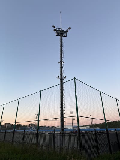 The image depicts a sports field or stadium at dusk. Dominating the foreground is a tall, industrial-looking light tower with multiple floodlights at its apex. The tower is positioned behind a chain-link fence with green metal posts, which encloses a rectangular area. This fence appears to be part of a larger sports facility, as evidenced by other similar light towers visible in the background, along with buildings and a treeline. The sky is clear and exhibits a gradient of light blue and a soft pinkish hue, indicative of sunset. In the immediate foreground, there is some overgrown grass and a dark, metal fence. The overall scene suggests an outdoor recreational or sporting venue preparing for evening use.