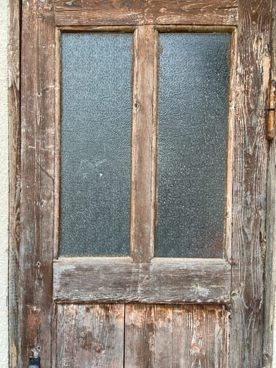 The image shows a close-up view of an old, weathered wooden door with frosted glass panels. The door is a dark brown color with significant signs of wear, including peeling paint, cracks, and a rough texture. It is divided into two sections by vertical wooden strips, and each section contains a frosted glass pane. The glass is opaque, obscuring any view beyond. On the right side of the door, a rusty metal hinge is visible. The door is set against a textured, light-colored wall. The overall impression is one of age and disuse. The location is given as Krasnomakskoye rural settlement, Ukraine. There are no people or animals visible in the image.