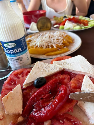 A detailed, high-angle shot captures a dining table set for a meal in Petrevene, Bulgaria. In the immediate foreground, a plate displays a large portion of a salad containing sliced red tomatoes, several irregular pieces of white feta cheese, some sprinkled with black pepper, and multiple large pieces of roasted red bell pepper. A single black olive is positioned on the cheese. A silver-bladed knife is partially visible on the right, resting on the food. Behind this plate, a tall white bottle of "Екомлеко Айран" (Ekomleko Ayran) with a blue label featuring Cyrillic text and a landscape illustration stands prominently. To the right of the bottle, a plate of golden-brown french fries is visible, generously topped with shredded white cheese. A silver cylindrical shaker is positioned to the left of the Ayran bottle. In the blurred background, the upper torsos of two individuals are seated at the table. One person on the left wears a light-colored top, while the other on the right wears a white V-neck top. Between them, a third plate of salad, containing sliced cucumbers, red vegetables, and white cheese, is visible. A red bowl and a gold-colored circular object are also present on the dark table surface.