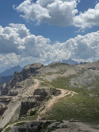 This is a daytime, vertical, eye-level, outdoor shot of the Tre Cime di Lavaredo in the Italian Dolomites, near Auronzo di Cadore. The image features a dramatic mountain landscape with rugged, rocky peaks and slopes covered in green grass and sparse vegetation. A well-worn dirt path winds its way across the mountainside, with several hikers visible on it, appearing to be walking or standing in groups. The sky is a vibrant blue, dotted with fluffy white cumulus clouds, suggesting fair weather. The terrain is characterized by steep inclines, sharp rock formations, and a sense of vastness.