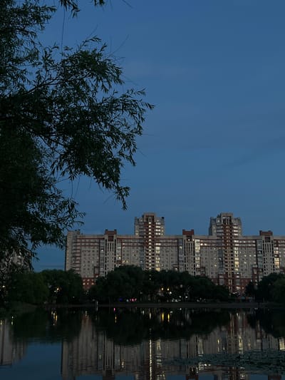 This image depicts a twilight scene in Rahya, Russia, with a large, modern apartment building reflected in a body of water. The building, constructed with a beige and red facade, features numerous illuminated windows, suggesting an evening or nighttime setting. Trees, some silhouetted against the dim sky and others reflected in the water, frame the scene. The water's surface is calm, creating a clear, inverted image of the building and trees. The overall mood is tranquil and serene, amplified by the twilight hues of the sky and the soft glow from the building's windows. The scene captures a moment of quiet beauty in an urban environment.