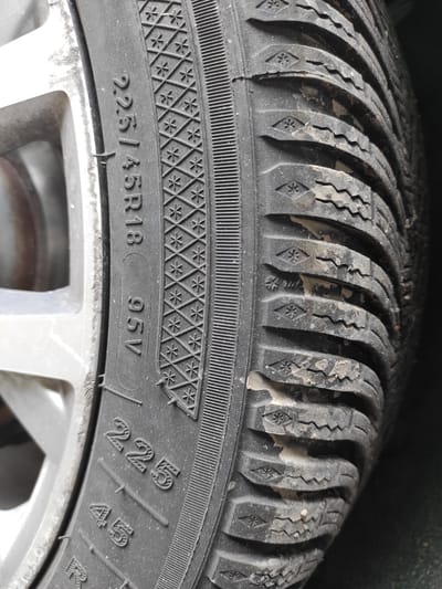 The image shows a close-up of a car tire, specifically the sidewall and a portion of the tread. The tire markings are clearly visible, indicating a size of "225/45R18 95V". The tread pattern suggests it is a winter tire, with small snowflake symbols embedded within the tire's sipes. There is some dirt and debris on the tire tread. A silver alloy wheel is partially visible on the left side of the image. The lighting suggests an outdoor environment, possibly a street or parking area. The location in Chaponost, France, is not discernible from the image itself, as there are no identifying landmarks or signage.