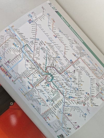 A printed "Regionaler Schienennetzplan" (Regional Rail Network Map) is displayed, oriented diagonally from the upper-left to the lower-right. The map features a white background with multi-colored lines representing various rail routes and black text identifying stations. The city of Niedernhausen, Germany, is visible as a station on the light blue S2 line, located in the central-left portion of the map. A section of an orange surface, possibly part of a train or station interior, is partially visible in the bottom-left corner, along with a small black circular object. No individuals are present.