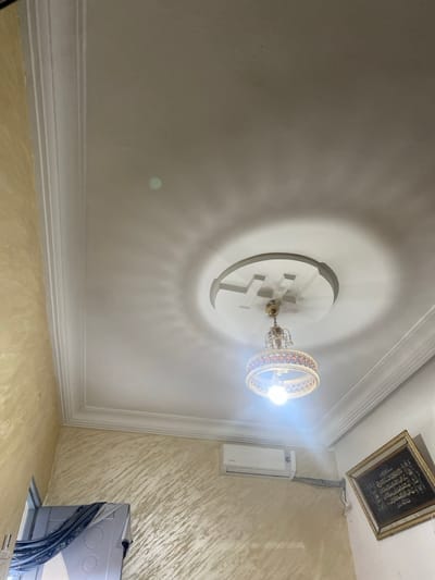 The image depicts an interior room in الغنادة, Tunisia. The white ceiling features decorative crown molding and a central circular medallion with a geometric, cross-like relief design. An illuminated, ornate chandelier with a circular, patterned shade and multiple bulbs hangs from the center of the ceiling, casting distinct radial shadows. The upper portions of the left and central walls display a textured, yellowish-beige finish. A white split-unit air conditioner is mounted high on the wall in the mid-right, with visible conduits extending horizontally. On the far right, a dark-framed artwork, presumed to be Islamic calligraphy, is mounted on a white wall. The lower-left corner shows a partial view of an opening or doorway, revealing a grey surface and a dark fabric draped over it. No people are visible.