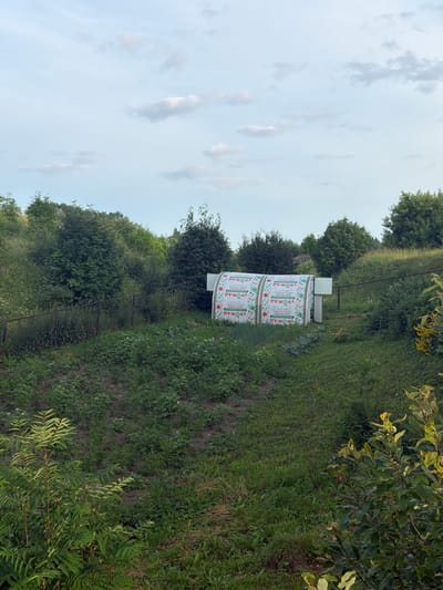 The image depicts an outdoor scene, likely in a rural or semi-rural setting in Cherdyn, Russia. In the foreground, there is a patch of ground covered in green grass and some low-lying vegetation. Beyond this, a raised area of soil is visible, also with green plants. A dark metal fence runs horizontally across the mid-ground, separating the foreground from a sloped embankment.
On the embankment, there is a tunnel-like structure, possibly a greenhouse or a temporary shelter, covered with a white material featuring a repeating pattern of red and green circular graphics and text. The text appears to be in Russian, with visible words like "УРОЖАЙ" (Urozhay), meaning "harvest." The structure has an open doorway with a white panel beside it.
Behind the tunnel structure, a dense line of trees and bushes forms a natural barrier. The background is dominated by a vast expanse of sky, with scattered white and grey clouds against a pale blue backdrop. The overall impression is a summer day in a landscape with cultivated areas and natural greenery. No people are visible in the image.