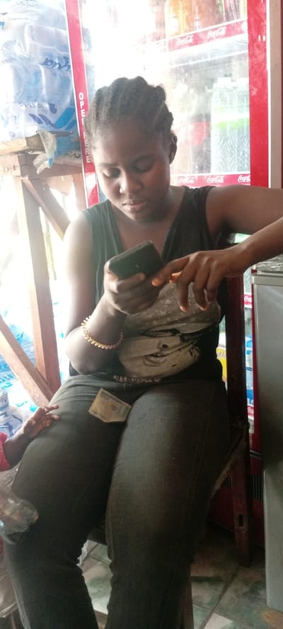 A woman is seated indoors, likely in a small store in Kwachiri, Nigeria. She's focused on a mobile phone she holds in her hands. She wears a dark tank top, jeans, and a gold bracelet. A banknote is visible on her lap. A refrigeration unit filled with Coca-Cola and bottled water is behind her. Plastic-wrapped water bottles are stacked to her left.