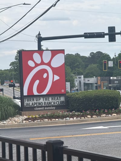 This image shows a Chick-fil-A sign in Stone Mountain, GA. The sign advertises a Jalapeno Ranch Club, and indicates that the restaurant is closed on Sunday. In the background, one can see traffic signals, trees, and other commercial buildings. The image was likely taken from an elevated position. The street is paved with double yellow lines, a white directional arrow, and a curb.
