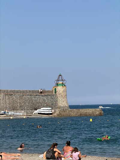 The image depicts a sunny day at a beach in Collioure, France, with a stone lighthouse and jetty dominating the background. Several people are visible enjoying the water and the shore. In the foreground, two women and a young girl sit on the pebble beach, facing the sea. One woman is wearing a striped swimsuit and the girl a pink t-shirt. Another woman in the foreground is partially visible, lying on her stomach.

Further out in the water, several individuals are swimming, including a man on a green inflatable raft. A couple is also visible swimming together. The sea is calm and blue, with a few yellow buoys marking a designated area.

The stone jetty stretches across the middle ground, topped with a unique lighthouse featuring a green cupola and metal framework. Several boats are moored along the jetty, and a few more are visible in the distance on the sea, including a white speedboat. The sky is clear and a pale blue. The overall scene conveys a relaxed and idyllic atmosphere of a summer day at the coast.