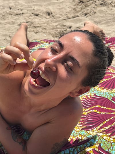 In this medium shot, a person with dark hair pulled back is lying on a sandy beach, with a colorful patterned fabric draped beneath them. The person is looking upwards with their mouth open, about to place a cherry into their mouth. Their eyes are closed, and their forehead is glistening with sweat, suggesting a warm, sunny day. The beach is visible in the background, with sand and a hint of water. The person has a nose ring and a tattoo on their arm, which is partially visible. The overall mood is relaxed and playful, possibly indicating a moment of enjoyment during a summer day at the beach in Gavà, Spain.