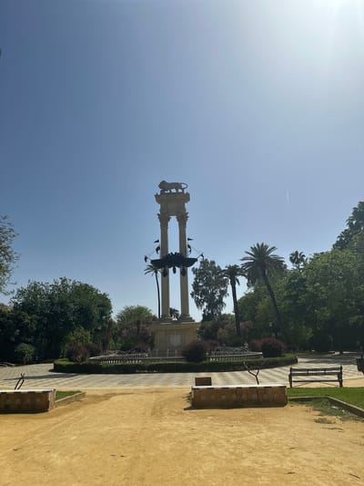 The image, correctly oriented as a landscape view, captures a sunlit park scene in what is identified as Sevilla, Spain. A prominent monument dominates the center, featuring two tall, light-colored classical pillars. Each pillar is crowned with a statue of a crouching lion, and between them stands an ornate, dark metal structure, possibly a multi-branched lamppost, adorned with bird-like decorative elements. The base of the monument also displays sculptural details, potentially a crest or relief.
The park is richly green with diverse vegetation, including several tall palm trees and dense broadleaf trees. A wide, sandy-colored dirt path occupies the right foreground and extends into the distance. Along this path, a long, built-in bench decorated with square, patterned tiles (azulejos) is visible. On the left side of the monument, a paved walkway with a geometric pattern runs parallel to the dirt path, separated by a low black metal fence and a strip of grass. A black metal park bench is situated on this paved area further back. In the background, a low, rounded hedge might encircle a fountain.
The sky is a clear, bright blue, indicating good weather. A strong sun flare is visible in the upper left quadrant, and a faint white contrail from an aircraft can be seen high in the sky. The scene is devoid of people or discernible activity, conveying a sense of tranquility. The bright illumination suggests it is daytime, likely mid-morning or afternoon. No visible text is legible on the monument or benches.