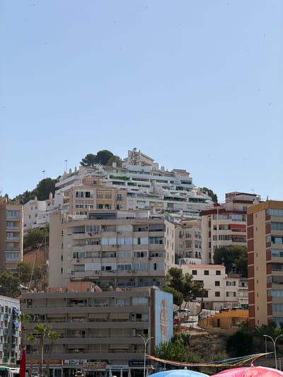 This media file shows a view of a densely populated urban area with multi-story buildings. The buildings are of various architectural styles and colors, with many featuring balconies and windows. Some buildings are modern, while others appear older. Green trees are interspersed among the buildings, suggesting a Mediterranean climate. In the foreground, there are some palm trees and colorful fabric banners, possibly indicating a festive event. A large mural of a vase is visible on one building.
The scene is set in an urban environment, likely a coastal town or city, given the architecture and vegetation. The bright, clear sky suggests it is daytime, likely mid-morning or afternoon, and the weather appears sunny and pleasant. There are no people clearly visible in the image.
Some visible text includes "Belber" on a red awning, and various signs on shops on the lower right side of the image, including "Cala Beach," "Waterfront," "EL TAPECITO," "PAPELERIA VELA," and "TALLER DE MODA." These texts, combined with the overall style of the buildings and vegetation, suggest the location is likely in Spain, as indicated by the location context. The overall impression is a vibrant, busy urban setting on a beautiful day.