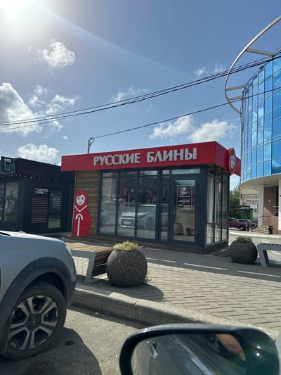 The image depicts a sunny daytime scene in Orsk, Russia, likely on a street or plaza. In the foreground, the left side of a silver SUV is visible, with its wheel and part of the fender in focus. To the right, a portion of a black car's side mirror reflects the surroundings.
The main focus of the image is a small retail kiosk with a bright red sign above it that reads "РУССКИЕ БЛИНЫ" (Russian Blini). The kiosk has large glass windows and a wooden facade. A large, stylized cutout of a traditional Russian nesting doll (Matryoshka) stands to the left of the kiosk entrance. Inside the kiosk, a person is partially visible behind the counter. Two round, stone planters with greenery sit on the paved walkway in front of the kiosk. Behind and to the right of the "Russian Blini" kiosk, a modern building with a blue glass facade and a curved metal structure is partially visible. Further to the right, a medical clinic sign "МЕДКИ" is present. Adjacent to the "Russian Blini" kiosk on the left is another smaller black kiosk with a "COFFEE LIKE" logo and its operating hours displayed on the window. The ground is paved with interlocking bricks, and a concrete curb separates the walkway from the street. The sky is blue with scattered white clouds and visible power lines.