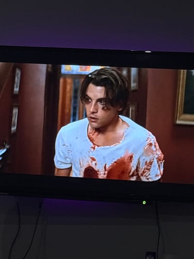 The image displays a photograph of a television screen showing an indoor scene. The specific location in the city of Ботево, Bulgaria, cannot be determined from the visual content presented.
On the television screen, a young adult male is depicted from the chest up, positioned slightly right of center. He has medium-length, wavy dark brown hair, fair skin, and dark eyes, looking intently to his left (viewer's right) with a serious or concerned expression. He is wearing a white t-shirt, heavily saturated with large, dark red stains consistent with blood, primarily across the chest, left shoulder, and stomach area, with smaller splatters on the right arm. The fabric of the t-shirt appears distressed, particularly around the neckline.
The background behind the male subject features an interior setting with dark reddish-brown walls and dark wooden architectural elements, possibly door frames or panels. On the left wall, two vertically oriented, rectangular framed objects are visible. On the right, a section of a framed painting with a gold-colored frame is partially discernible. Further back, a decorative glass or stained-glass window with geometric patterns in shades of yellow and blue is visible.
Below the television screen, a black flat-screen TV bezel is visible, featuring a small green indicator light on its lower right side, indicating power. Below the bezel, dark cables descend against a plain, light-colored wall. A subtle purple ambient light glow is present on the wall below the television. No direct interactions or specific actions other than the male subject's focused gaze are observable within the depicted scene on the screen. The presence of significant bloodstains implies prior violent activity or injury.