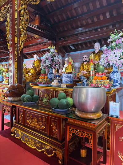 This photograph depicts a richly decorated altar in what appears to be a temple in Diên Khánh, Vietnam. The structure is predominantly constructed from dark wood and is ornately adorned with gold-colored carvings. Arranged on the altar are several gilded statues, possibly of Buddhist figures. Between the statues are various offerings, including vases of flowers (lilies), porcelain vases in blue and white, and an assortment of fruits like oranges, dragon fruit, and pomelos, some of which are grouped in bowls. A large silver vessel also sits on the altar, alongside a coiled golden decorative item. The temple's interior features a dark wooden ceiling with exposed beams and gold details. A QR code is visible on a sign attached to the altar. The overall scene is one of reverence and cultural significance.