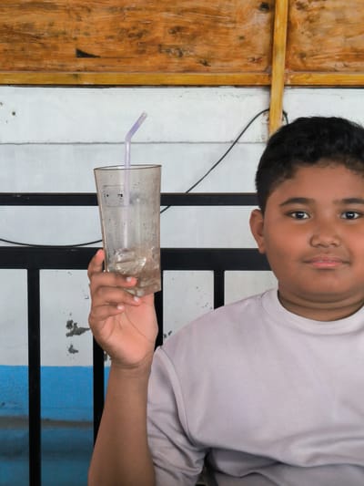 A boy stands in front of a fence with black vertical bars against a white wall. The lower portion of the wall is blue. Behind the wall is a wooden structure with varying shades of brown. The boy has dark hair and a light brown complexion. He wears a light-colored shirt. He holds a transparent glass filled with ice cubes and a purple straw. The photo was taken in the city of Matang, Indonesia.
