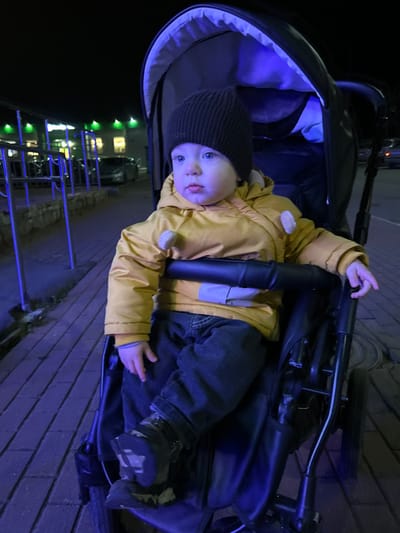 A young child, likely a toddler, is seated upright in a dark-colored stroller at night. The child wears a brown knitted beanie, a yellow padded winter jacket with light-colored decorative elements, dark blue jeans, and dark shoes with patterned soles. The child has light skin and blue eyes, gazing slightly to the left with a neutral expression. A strong blue light illuminates the interior of the stroller's canopy and the lower portion of the child and stroller. The ground is paved with reddish-brown bricks. To the left, a light-blue illuminated railing or barrier runs parallel to a building with illuminated windows and a series of green lights installed along its roofline. Multiple vehicles are visible in the background on what appears to be a street or parking area. The scene depicts an outdoor urban setting in Voskresensk, Russia.