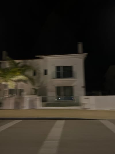 A blurry, sideways nighttime view reveals a white building with dark-windowed rectangular sections. A palm tree is partially visible near the building's base. A gray surface with blurred white lines occupies the right side of the frame. The left side is dominated by black.