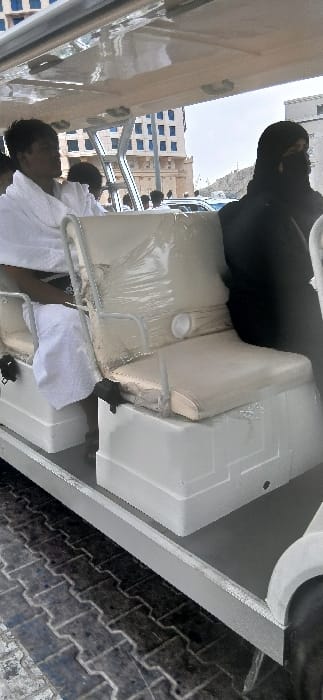 The image depicts a daytime scene in Makkah Al Mukarramah, Saudi Arabia, featuring an open-sided electric transport cart on a paved surface. Inside the white-framed cart, a male individual wearing white Ihram garments is seated on the left, facing forward. To the right, a female individual dressed in a black abaya and niqab is seated, with only her eyes visible. An empty, light-colored upholstered seat, partially covered in clear plastic wrap, separates the two main visible passengers. The cart's roof is white and translucent. The ground beneath the cart is paved with dark grey interlocking paver blocks. In the background, several multi-story buildings, consistent with urban pilgrim accommodation, are visible under a bright sky. One prominent building displays blue-framed windows. A rocky, arid hill is visible on the far right background, and other vehicles and pedestrians are present in the distance. The individuals in the cart are static, appearing to be passengers.