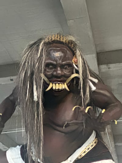 An imposing Balinese Ogoh-ogoh statue is depicted in a full-frame, eye-level shot, likely located under a concrete overhang or structure in Celuk, Indonesia. The statue features dark brown skin, a menacing expression with wide-open eyes, and a grimacing mouth revealing prominent yellow human-like teeth on the lower jaw, flanked by two upward-pointing white fangs. Two large, curved, ivory-colored fangs protrude downwards from the upper jaw. The figure has long, disheveled dark hair with lighter streaks, a golden snake-like crown, and a red and gold ornament near its right ear. Its muscular body is minimally clothed, with a white fabric draped around the waist and a golden patterned sash or belt visible. The statue's left hand, featuring long, yellow, claw-like nails, is extended forward, conveying a reaching or grasping posture. The background consists of undetailed gray concrete surfaces and beams, indicating an artificial overhead structure.