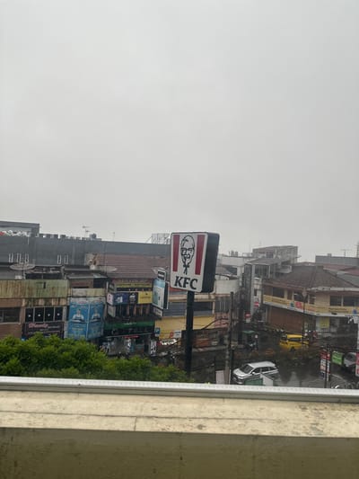 This image captures an urban street scene in Sukabumi, Indonesia, viewed from a slightly elevated perspective, likely a window or balcony. The sky is overcast and bright, suggesting a cloudy day with diffuse lighting, and some wet surfaces on the street indicate recent rain.
The most prominent subject is a large, square KFC sign, featuring the white "KFC" text and Colonel Sanders' face on a black background, mounted on a pole. Below it, several other commercial signs adorn the multi-story buildings that line the busy street. Visible brand names and texts include "realme", "OPPO", "PULSA", and what appears to be "RICHARD HANS". Other signs show images and text related to "MS GLOW" and "SKINCARE".
The buildings themselves are varied in style and height, some featuring traditional tiled roofs and others appearing more modern with concrete facades. Numerous overhead power and utility lines crisscross the scene.
On the street below, several vehicles are visible, including a white car (possibly a compact hatchback), a yellow car (perhaps a taxi), and a green bus. Lush green foliage, possibly trees or bushes, lines the lower right side of the frame, partially obscuring the street. The right edge of the image is framed by a light-colored concrete or stucco wall, reinforcing the idea of a view from an interior space. No specific activity or event is taking place; it appears to be a typical bustling street during daytime.