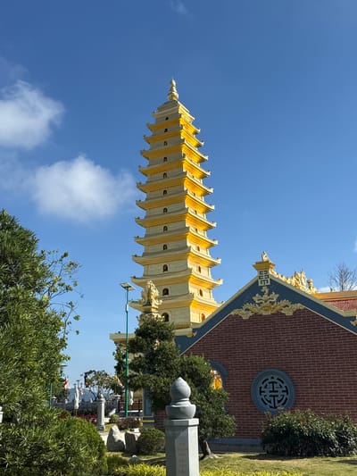 The image shows a multi-tiered, yellow and white pagoda against a clear blue sky with some clouds. The pagoda is situated next to a red brick building with a round decorative window and gold accents. In the foreground, there are trees and vegetation, and a gray concrete structure resembling a memorial or marker. The setting appears to be a temple or park, likely located in Diên Khánh, Vietnam. The photograph is taken from a low angle, emphasizing the height of the pagoda.