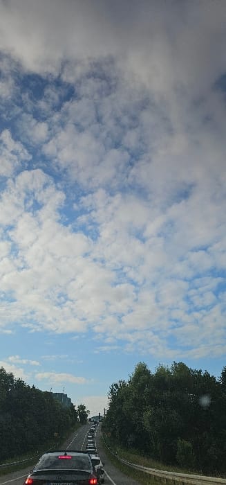 The image captures a traffic jam on a two-lane road in Hermsdorf, Germany, from a vehicle's perspective. The upper two-thirds of the frame show a sky featuring scattered white cumulus clouds against a blue background, transitioning to a denser, grey cloud layer overhead. The lower third depicts the road, lined on both sides by dense green deciduous trees and bushes.

A dark-colored Audi sedan, bearing the German license plate "WH T 866R" with illuminated red taillights, is prominently positioned in the immediate foreground, indicating the vehicle is stationary or moving slowly. A continuous line of additional vehicles extends into the distant background, signifying significant traffic congestion on the ascending road. A light-colored guardrail is visible along the right side of the road, parallel to the vegetation. On the left, in the mid-distance, a multi-story, light-colored building with associated infrastructure is partially visible above the tree line. No individuals are discernible. A small light flare is present in the mid-right of the image.