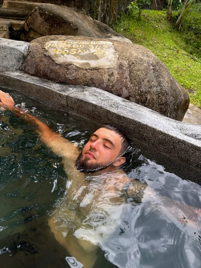 A man with dark hair and a beard is partially submerged in a stone hot spring, his eyes closed and his right arm extended above the water. He has tattoos on his arm and chest that are visible through the water. The hot spring is surrounded by large rocks and lush green vegetation. In the background, there are stone steps leading up a grassy slope. A weathered stone plaque is embedded in a large rock near the edge of the hot spring. The plaque appears to have yellow lettering, but the text is difficult to decipher. The setting is likely an outdoor natural area in or around Khanh Vinh, Vietnam, known for its hot springs.