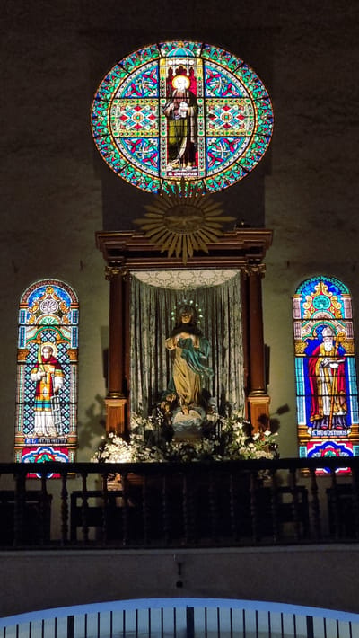 The image shows the interior of a dimly lit church, likely in Cadaqués, Spain. The scene is dominated by religious iconography and architectural elements.
In the center, a statue of a robed figure, possibly a saint or religious icon, is displayed in an alcove. The statue is adorned with a halo and surrounded by a profusion of white flowers. Behind the statue, a draped curtain creates a dramatic backdrop. To the left of the statue, a large, circular stained-glass window features intricate geometric patterns and a central depiction of a person in religious attire. Adjacent to this window is a golden, sunburst-like emblem.
Above and below the central display are two rectangular stained-glass windows. The upper window depicts two figures in religious vestments, with visible text on the right side that reads "S. AUGUSTINUS". The lower window also features a religious figure, though the text is less legible.
The overall atmosphere is solemn and reverent, consistent with a religious setting. The lighting suggests it is either evening or a time when artificial illumination is needed within the church. The architectural style and the presence of stained-glass windows are characteristic of European churches, and the specific mention of "S. AUGUSTINUS" may refer to Saint Augustine, a prominent figure in Christian theology and history.