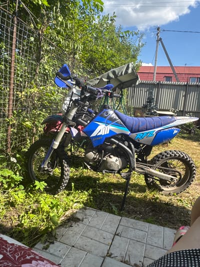 In the city of Gal’chino, Russia, a blue and white dirt bike is parked on a dirt and grassy area next to a chain-link fence and overgrown green foliage. The bike is positioned at an angle, with its front wheel slightly turned to the left. A green tarp or canopy is draped over the handlebars and front of the bike. In the background, a corrugated metal fence separates the yard from a residential building with a red roof. A utility pole with wires stands against a blue sky with white clouds. The ground in the foreground is partially covered with light-colored paving stones. A person's legs, wearing a black and white polka dot skirt and pink sandals, are visible in the lower right corner of the image.