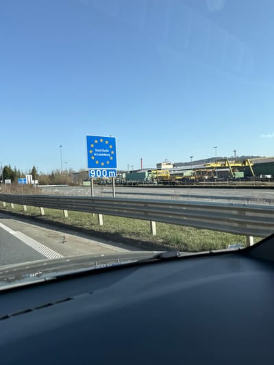 The image displays a daytime scene from the interior of a vehicle, looking out through the windshield. A blue rectangular road sign, bearing the European Union circle of stars at the top, indicates "Grand Duché de Luxembourg" in white text, followed by "900m". This sign is positioned on the right side of a multi-lane road, adjacent to a grey metal guardrail. The road itself features white dashed lines and appears to curve to the left. In the background, beyond the road and the sign, an industrial landscape is visible, characterized by large, low-rise buildings, including some with green siding, and prominent yellow gantry-like structures. Piles of dark material are present on the ground within the industrial area. A tall, slender red-and-white striped structure, possibly a chimney or tower, stands to the left of the main industrial complex. Utility poles and lines are scattered across the horizon, which is framed by a clear, bright blue sky. Sparse trees are visible on the far left horizon. The foreground shows the dark dashboard of the vehicle and the bottom edge of the windshield. The scene depicts the approach to the Grand Duchy of Luxembourg border from Aubange, Belgium.