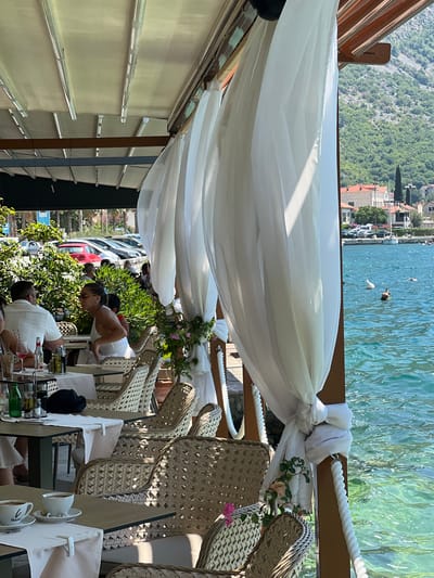 The image depicts a waterside restaurant in Risan, Montenegro, likely during the daytime. White, sheer curtains are draped from a retractable awning, creating a semi-enclosed dining area. Several tables are set with white tablecloths and wicker chairs, some of which are occupied by diners. In the foreground, two tables are visible, one with cups of coffee and the other with drinks and condiments. In the mid-ground, several individuals are seated at tables, including a woman in a white dress and sunglasses and a man in a white shirt. Beyond the restaurant seating, a body of turquoise water is visible, with a few people swimming or wading. In the background, a hillside covered with lush greenery and dotted with red-roofed buildings rises steeply. The overall scene suggests a relaxing and pleasant dining experience with a scenic view.