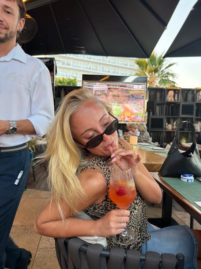 A blonde woman with sunglasses on her nose drinks from a cocktail with a straw. She is wearing a leopard-print top and denim shorts, and her left arm is bent, supporting her head. She is seated at a table with a glass of orange cocktail, a black handbag, and a small blue container. To her left, a man in a white shirt and dark pants stands, partially visible. Behind them, a large television screen displays a sporting event. In the background, there is a building with balconies, a palm tree, and shelves filled with decorative items. The scene appears to be an outdoor dining area at a restaurant in Calvià, Spain, with tables and seating visible.
