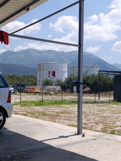 This media file depicts an outdoor industrial or commercial setting under a bright, sunny sky with scattered clouds. The scene is dominated by large, white cylindrical storage tanks, one of which features a prominent red and white "EKO" logo. In the foreground, a portion of a silver car is visible, parked on a concrete surface with a drainage grate. A red cloth is hanging from a metal structure overhead, suggesting a car wash or maintenance area.
Behind the car and the storage tanks, a chain-link fence encloses the area. Through the fence, various vehicles are visible, including trucks, an orange van, and a yellow excavator. Trees and foliage form a backdrop to the industrial elements, and a mountain range looms in the distance under the blue sky.
The lighting suggests it is daytime, likely mid-morning or afternoon given the shadows. The overall impression is one of an operational industrial site, possibly related to fuel storage or distribution, given the EKO branding and the presence of large tanks and heavy machinery. There is no visible indication of people or any specific activity beyond the static presence of vehicles and infrastructure. The location is identified as Mrčevac, Montenegro.