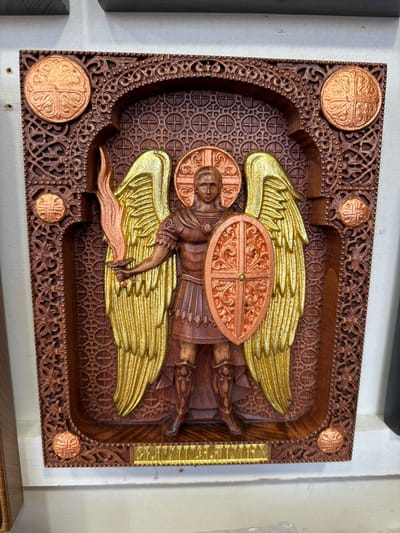 A carved wooden icon depicting Saint Michael the Archangel is displayed on a white surface, likely a shelf or table. The icon is richly detailed, with Saint Michael shown in armor, holding a fiery sword and a shield. His wings are large and outstretched, rendered in gold leaf. The surrounding frame of the icon is intricately carved with decorative patterns and features four circular copper accents, each with a cross design. Below the figure, a golden plaque with inscribed text is visible. The background within the icon features a repeating pattern of circles containing crosses. The overall aesthetic suggests a religious artifact. The location is specified as Yalta, Ukraine.