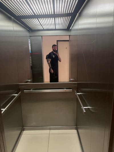 A man stands in an elevator, looking into the reflection of a mirror panel. The elevator walls are metallic. One wall features a grid-like pattern of holes. Buttons for different floors are visible near the mirror panel. The location is Cordoba, Argentina.