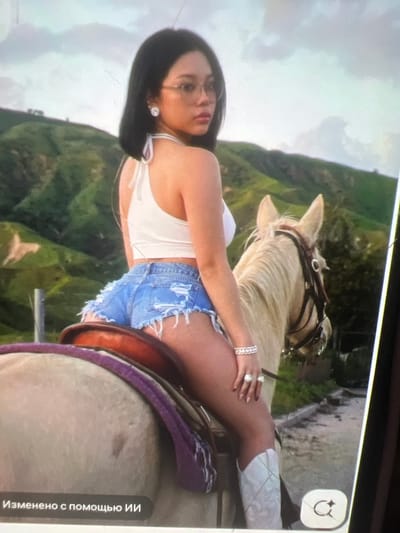A young woman with dark hair and glasses sits atop a pale horse in a hilly, green landscape. She is wearing a white halter top, ripped denim shorts, and white cowboy boots. She is adorned with jewelry, including earrings, a necklace, a bracelet, and rings. The horse is equipped with a saddle and bridle. The background consists of rolling hills covered in vegetation, with a cloudy sky above. The image appears to be taken outdoors, likely in a rural or natural setting. The location is identified as Kara-Dıykan, Kyrgyzstan.