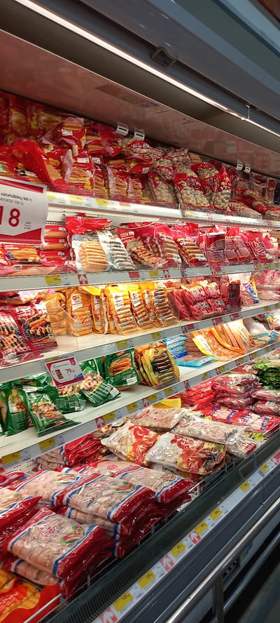 This media file depicts an overhead view of a supermarket or grocery store freezer aisle in Moo 1, Thailand. The primary subjects are numerous packaged food items, predominantly different varieties of sausages, hot dogs, and other processed meat or fish products. These items are displayed within long, multi-tiered refrigerated or freezer cases.

The activity taking place is the commercial display of food products for sale. The items are organized and stacked in individual clear plastic packages, many featuring vibrant red, green, or yellow branding and labels. The display cases are well-lit, highlighting the products.

Notable details include the extensive variety of similar products, suggesting a specialized section for these items. Several price tags are visible along the edges of the display cases. Visible text includes prices such as "129", "299", "189", "205", and "79.00". A larger red and white sign prominently features a large "8" and "79.00". One product label clearly shows the Thai text "แฟรงค์แม่ชีร์หนู 500 ก." (Frankfurter 500 g.) and the English text "NKFURTER 500 G.". No people are visible in the image, and the time of day is not discernible.