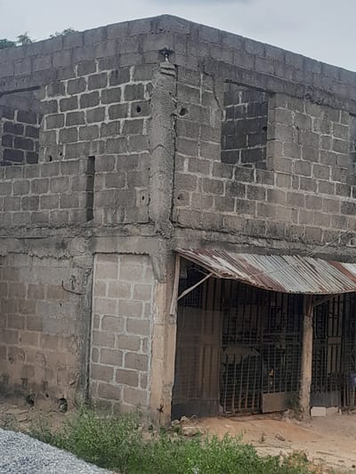 The scene depicts a two-story concrete block structure in Ifo, Nigeria, under an overcast sky. The building is constructed from unrendered grey concrete blocks, exhibiting an unfinished state or significant disrepair. The upper floor features multiple rectangular openings consistent with unglazed windows. A prominent, roughly finished horizontal concrete beam separates the ground and upper levels. The lower left side of the building contains a blocked-up opening and a doorway filled with concrete blocks. Extending from the lower right side is a rusted, corrugated metal awning, supported by unpainted wooden posts. Beneath this awning, multiple dark metal security grates or gates are visible, suggesting closed entrances or storefronts. The ground directly in front of the structure is unpaved earth, covered with patches of green weeds and scattered debris. A pile of light-colored gravel occupies the lower-left foreground. No individuals are present or engaged in any discernible activity within the frame.