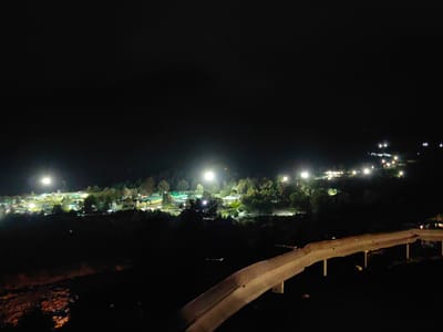 The image shows a nighttime view of a settlement with buildings and trees illuminated by artificial lights. In the foreground, a concrete barrier with supporting posts is visible, suggesting it is part of a road or an elevated structure. The background features a landscape with a dense arrangement of buildings that are lit from within and by streetlights, indicating a populated area. Trees are scattered throughout the settlement and the surrounding areas. A bright spotlight illuminates a central area of the settlement. The sky is dark, suggesting it is nighttime. The overall scene depicts a tranquil, illuminated settlement at night, potentially nestled within a natural landscape. The specific location in Palchan, India, is suggested by the request, but this cannot be confirmed visually from the image alone.