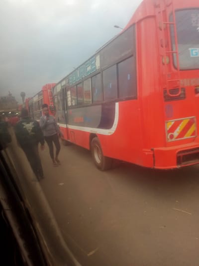 A low-angle, medium shot captures a street scene in Hospital Ward, Kenya, where a line of red and grey public transport buses are parked. The buses appear to be in a city setting, with a cloudy sky overhead. The bus in the foreground, predominantly red, has "SOURCE" printed in teal on its side. Two individuals are visible on the pavement beside the bus: a man in a dark jacket with a design on the back, and a woman in a grey top and dark leggings, holding a phone. The bus has a ladder attached to its side, suggesting maintenance or access to the roof. Another bus, visible behind the first, shares the red and grey color scheme. The road surface is asphalt, with some debris scattered on it. The overall atmosphere is subdued, possibly due to the overcast weather.