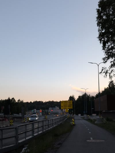 The image shows a street scene in Vantaa, Finland, likely near an industrial or commercial area given the signage and presence of what appears to be a commercial building. The sky is a soft gradient of blue and light orange, indicating either dawn or dusk. A street with a pedestrian path runs diagonally from the lower right to the mid-left of the frame. A silver car is visible on the road, traveling away from the viewer. Construction barriers and temporary traffic signs are present, suggesting ongoing road work. A yellow directional sign is visible on the left, pointing to "TIKKURILA DICKURSBY", "PORTTIPUISTO", and "IKEA" with arrows indicating their directions. Two streetlights are present, one in the foreground and another further in the background. The area is bordered by trees on the left and right, and a dark, modern-looking building is situated on the right side of the frame.