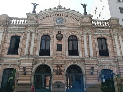 The image shows the facade of Casa de Correos y Telegrafos, in Lima, Peru, now La Casa de la Gastronomia Peruana. The building is a peach color, ornate, with statues of winged figures at the top of the facade. There are 2 stories, each with arched windows and arched doorways at the bottom. A sign above the second story windows reads "Casa de Correos y Telegrafos," with a clock above it. Below, above the doorways, a sign reads "La Casa de la Gastronomia Peruana." A person stands in front of one of the doors at the bottom. The sky is overcast. A light-colored building is partially visible on the right.