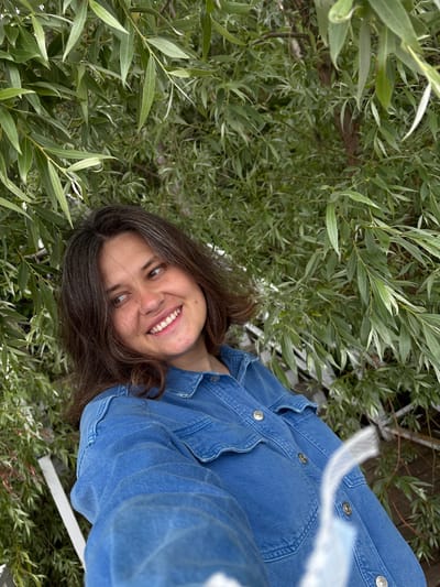 A young woman with brown hair is looking to her right and smiling. She is wearing a blue denim jacket and is surrounded by green foliage. The background appears to be a natural setting, possibly a park or a garden. There are no visible structures or landmarks that would suggest a specific location within Kostovaty, Russia.
